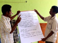 Two men hold up and explain financial information on a big sheet of paper.